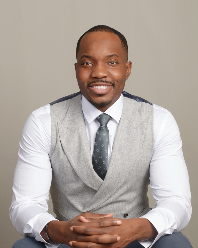 Dr. Bryant Ukaigwe seated in a gray vest and tie, hands clasped, smiling